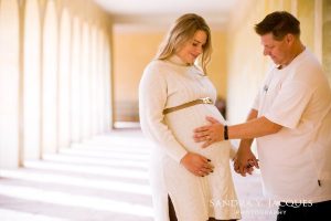 Ein Mann berührt den Babybauch seiner Frau. Beide stehen in einem hellen Säulengang ©sandra-jacques